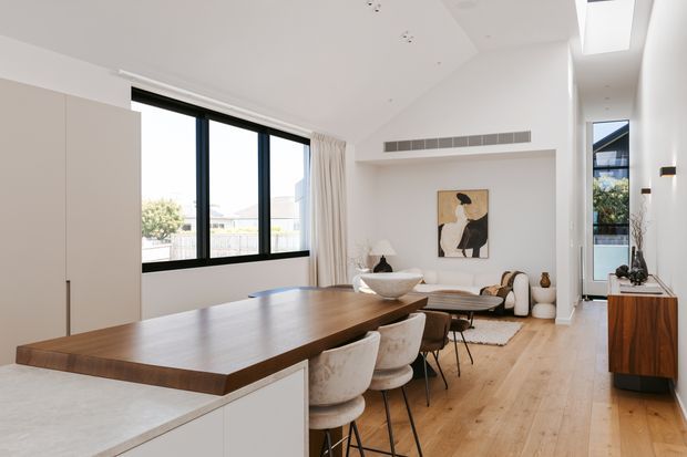 Bright open-plan contemporary interior combining a kitchen island and dining area with an adjoining living room. The space features pale timber flooring, a large dark timber island benchtop with bar seating, white cabinetry, black-framed glazing, a skylight, neutral furnishings, and restrained artwork. A linear air-conditioning grille and a hallway or secondary space are visible beyond the living area.
