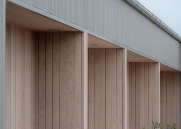 Close-up exterior architectural view of a modern building facade, featuring pale vertical timber cladding recessed beneath a deep overhanging roof or canopy. White corrugated metal cladding forms the upper facade, with exposed roof drainage piping along the edge. The composition emphasizes repetitive bays, clean lines, and restrained natural materials.