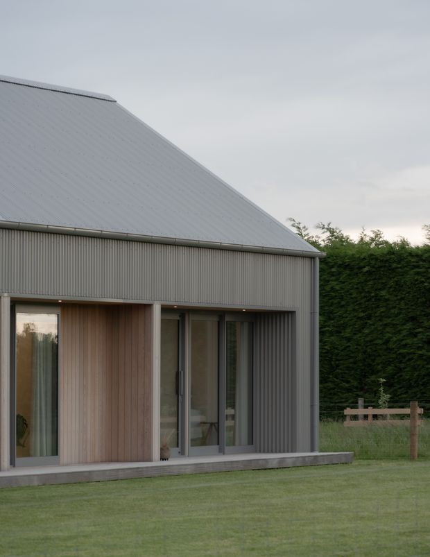 Exterior view of a compact contemporary house with a steep gabled corrugated metal roof and matching vertical ribbed metal cladding. Full-height glazed doors and windows are set within a warm timber-lined recessed entry or porch. The house sits beside a neatly mown lawn, dense hedging, and a simple timber fence under an overcast sky.