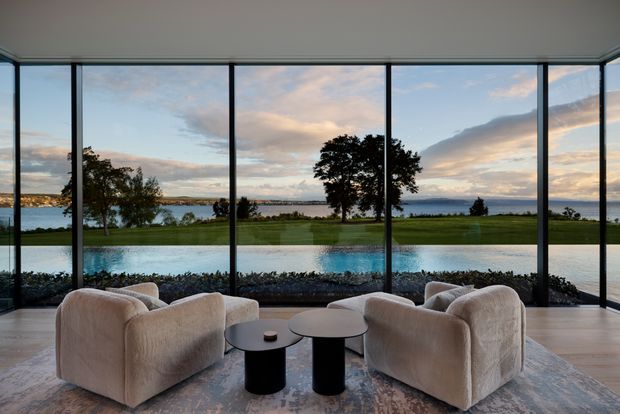 A refined living room with four curved, upholstered lounge chairs arranged around two small round coffee tables. Full-height glazing spans the room, opening views across a lawn, reflecting pool or water feature, and a broad waterfront landscape. The interior has a calm, neutral palette with pale timber or stone flooring, a large muted rug, and a minimalist contemporary fit-out.