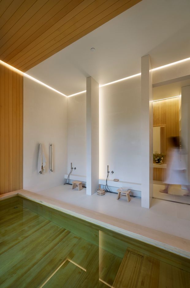 A serene indoor spa and pool area with a green-tinted rectangular plunge pool in the foreground. The minimalist space has pale plaster walls, slim white structural columns, warm timber wall and ceiling panelling, concealed linear LED lighting, and a built-in bathing or shower zone with stools and fixtures. A mirrored or glazed opening reflects the timber-lined adjacent area and a blurred person.