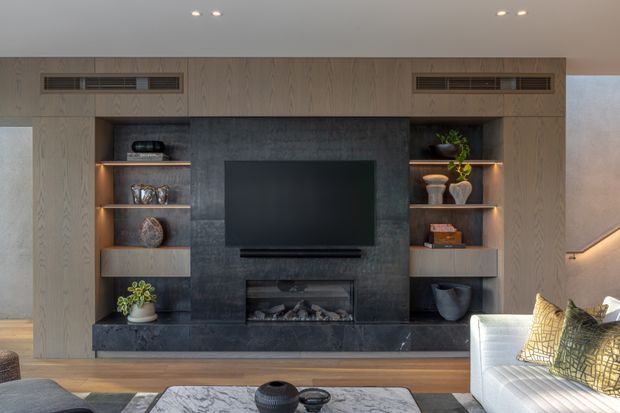 A contemporary living room media wall with extensive light timber built-in cabinetry, open display shelving, a dark stone feature wall, wall-mounted television and integrated fireplace. A pale sofa and marble-look coffee table occupy the foreground, with warm shelf lighting and timber flooring creating a refined, cozy atmosphere.