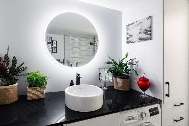 A contemporary bathroom vanity with a round backlit mirror above a white vessel basin on a glossy black benchtop. White built-in cabinetry includes a front-loading washing machine, while potted plants, a red glass vessel, and monochrome wall art add decorative accents. A tiled shower is visible in the mirror reflection.