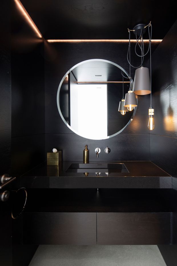 A compact, dramatically styled bathroom with black timber-look wall panelling and black cabinetry, a dark rectangular vanity with integrated basin, wall-mounted brass fixtures, a large round mirror, and sculptural hanging pendant lights. Warm concealed LED lighting and brass accents contrast with the predominantly black interior, while a light grey floor softens the space.