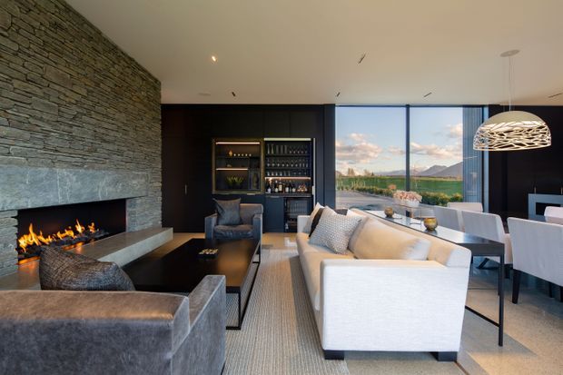 A sophisticated open-plan living and dining room with a long stacked-stone fireplace wall and linear gas fire, dark built-in bar cabinetry, contemporary upholstered seating, a dining table, and a sculptural pendant light. Full-height glazing opens to expansive mountain and water views, creating a refined indoor-outdoor connection.