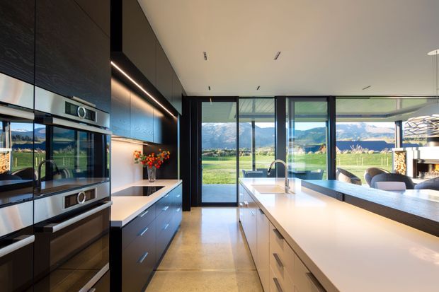 A bright contemporary kitchen with a large central island, integrated stainless-steel appliances, dark full-height cabinetry, and expansive black-framed glazing opening to a mountain and rural landscape. The island includes a sink and generous white benchtop, while a dining area is visible beyond.