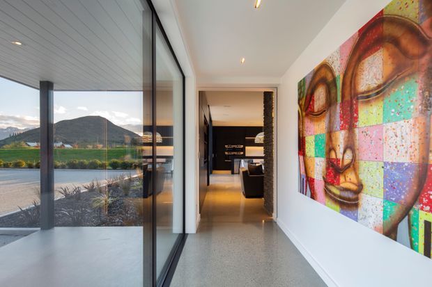Contemporary residential hallway with polished grey concrete or terrazzo flooring, white walls and ceiling, and full-height black-framed glazing overlooking a landscaped exterior and mountain setting. A large colourful abstract artwork provides a bold feature along the right wall, while a dark furnished living area is visible at the end of the corridor.