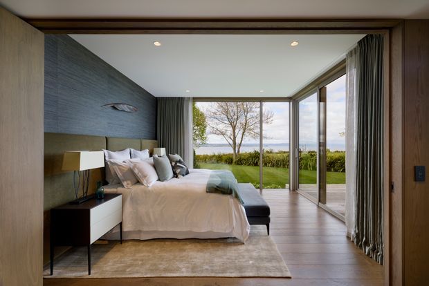 A contemporary bedroom viewed through a timber-framed opening, featuring a low upholstered bed with white bedding, dark bedside furniture, a textured dark feature wall, timber flooring, full-height glazing, and curtains opening to a landscaped lawn with a distant water view.