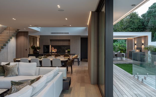 Contemporary open-plan living and dining area with a white sectional sofa, timber flooring and wall panelling, a dining table, and a dark built-in kitchen or joinery wall. A stairway is visible at left. Full-height glazing opens onto a timber deck and landscaped courtyard with a covered outdoor dining area and glass pool or terrace balustrade.