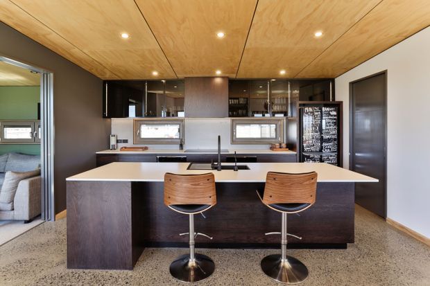Contemporary kitchen viewed directly from the living area, featuring a large dark timber island with a pale benchtop, two bar stools, integrated sink and tapware, dark cabinetry, mirrored splashback cabinetry, and a warm plywood-lined ceiling. A bedroom or living space is visible through the opening on the left.