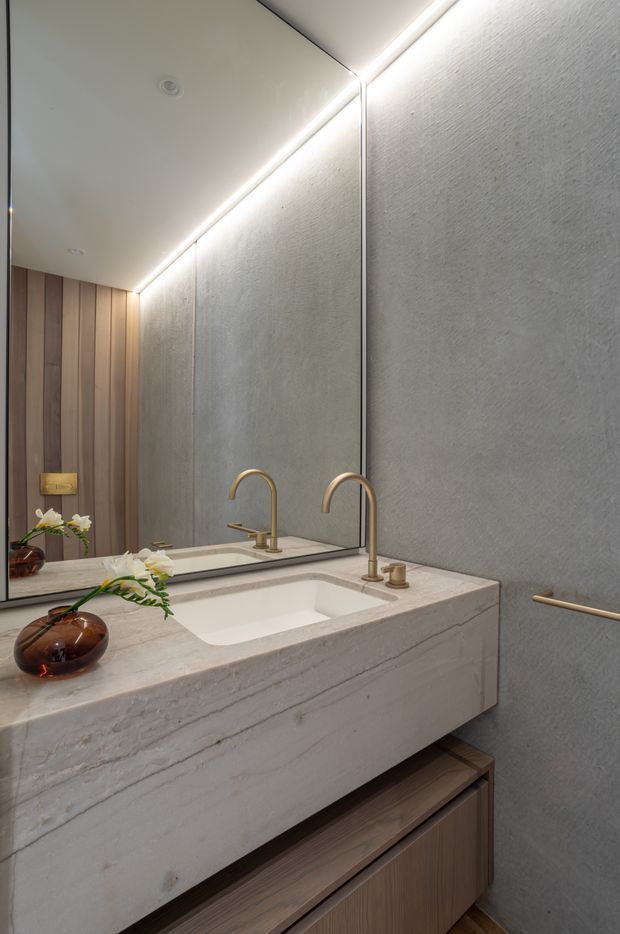 A refined contemporary bathroom vanity with a large frameless mirror, integrated warm LED lighting, a pale textured stone benchtop and basin, brushed brass curved tapware, and dark timber veneer cabinetry. Vertical timber wall panelling and subtle decorative styling add warmth to the otherwise neutral space.