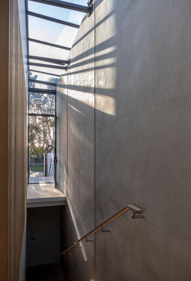 A narrow, tall stairwell or passage with a descending stair and a simple metal handrail mounted to a textured plaster wall. Translucent overhead glazing and side glazing bring in strong natural light, casting linear shadows across the wall. Timber cladding or panelling is visible along the left side, with an exterior deck and garden glimpsed beyond the glazed opening.