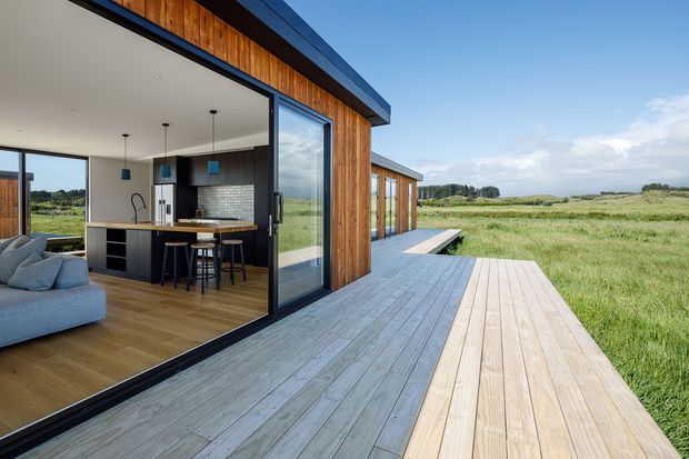 Contemporary single-storey home opening onto a broad timber deck, with a compact open-plan kitchen and living area visible through large black-framed glazing. The exterior uses vertical timber cladding, while the deck extends into an expansive rural grassland setting. The interior features dark kitchen cabinetry, timber flooring, pendant lights and exposed white ceilings.