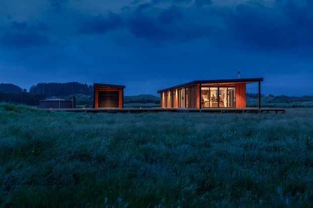 A low, single-storey contemporary house set in an expansive grassy landscape at dusk. The long linear building features warm timber cladding, extensive floor-to-ceiling glazing, a dark flat roof, and a separate timber-clad garage or outbuilding connected by a low platform. Interior lighting glows through the windows, creating a strong contrast with the deep blue evening sky.