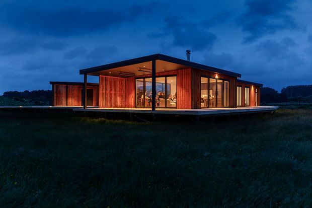 A low, single-storey modern pavilion-style house photographed at dusk, set in an open grassy landscape. The building has a broad flat roof with deep overhangs, extensive floor-to-ceiling glazing, dark structural framing, and warm timber-clad walls glowing from interior lighting. A raised deck-like platform surrounds the house, with an open-plan living area visible through the glass.