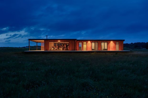 A long, low-profile single-storey dwelling photographed at dusk, set within an open grassy landscape. The building has warm timber-clad walls, a dark shallow roof, extensive glazing, and a covered outdoor terrace or veranda at the left. Interior lighting creates a warm glow against the deep blue evening sky.