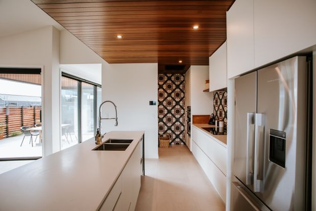 Bright contemporary kitchen with a large white island, integrated sink and faucet, white cabinetry, stainless-steel refrigerator, timber slatted ceiling, and a patterned tiled feature wall. Full-height glazing opens toward an outdoor dining area and deck.