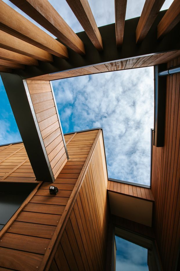 Upward-looking architectural photograph of a narrow courtyard or atrium framed by warm timber-clad walls, exposed timber roof beams, and dark structural elements. A large open skylit void reveals blue sky and clouds, emphasizing the vertical geometry and layered roof structure.