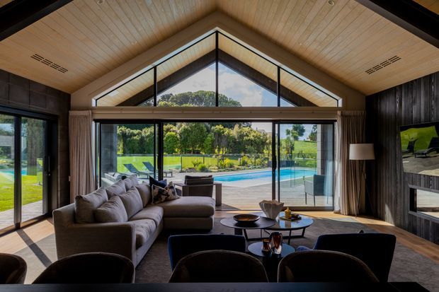 A spacious contemporary living room with a dramatic gabled, wood-lined ceiling and full-height glazing overlooking a swimming pool and landscaped grounds. The room features a large sectional sofa, lounge chairs, a low coffee table, dark vertical wall cladding, and a fireplace/media wall. Strong indoor-outdoor connection and abundant natural light define the space.