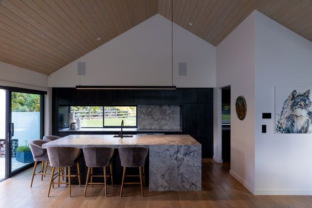 Contemporary open-plan kitchen with a large stone island and four upholstered bar stools, set beneath a warm timber-lined vaulted ceiling. Dark cabinetry and a veined stone splashback contrast with white walls, timber flooring, expansive glazing, and minimalist pendant lighting. A hallway and artwork are visible beyond the kitchen.
