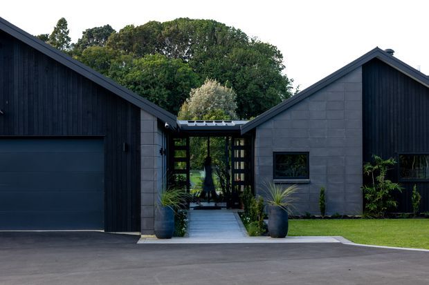 Exterior view of a contemporary single-storey residence composed of multiple dark gabled volumes around a glazed central entry courtyard. The facade combines near-black vertical timber cladding with grey masonry panels, dark-framed glazing, a broad garage door, and a paved approach with large planters and landscaping.