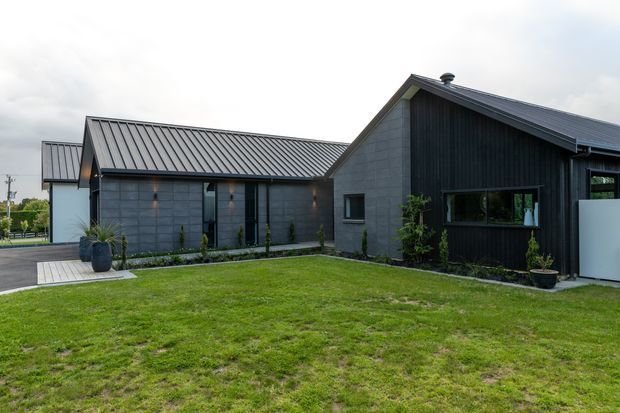 Exterior view of a contemporary single-storey house arranged around a broad grass lawn. The home has intersecting pitched roof volumes with dark metal roofing, charcoal vertical cladding, grey masonry wall sections, dark-framed windows, and a simple paved entry path. Minimal landscaping and restrained dark tones give the facade a modern, understated appearance.