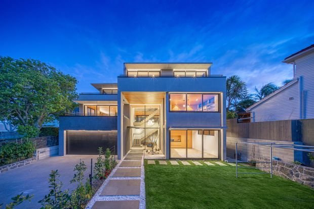 Twilight exterior view of a contemporary multi-level residence with a boxy flat-roof form, dark rendered facade, extensive glazing, warm timber soffits, balconies, and a central glazed entry stairwell. The home fronts a landscaped lawn with a paved path, driveway, planted garden beds, and a rendered boundary wall.