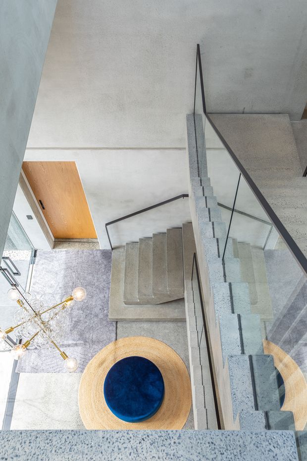 Looking down from an upper landing into a contemporary stairwell, with a grey terrazzo or concrete staircase, dark-framed glass balustrade, and pale concrete walls and floors. A circular woven rug with a deep blue round ottoman, grey rug, and sculptural brass light fixture are visible below.