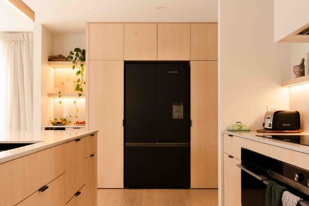 Warm contemporary kitchen featuring light timber cabinetry, a large central black refrigerator integrated into full-height joinery, pale benchtops, open display shelving, and a compact island. Soft concealed and under-shelf lighting creates an inviting atmosphere, with a toaster and oven visible on the right and a curtained window on the left.