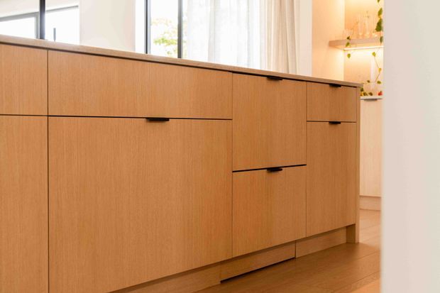 Close-up interior view of a long built-in timber-grain cabinetry unit with flat slab doors and drawers, recessed dark pulls, and a dark countertop. The warm wood joinery dominates the foreground, with tall windows, sheer curtains, open shelving, and a light-filled room visible behind it.