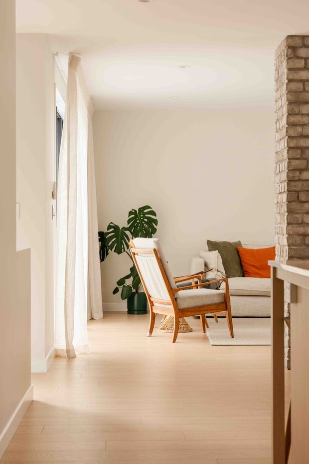 A bright, warm-toned living room viewed from an adjoining hallway or entry. The room has pale timber flooring, off-white walls and ceiling, sheer full-height curtains, a light upholstered sofa with olive and orange cushions, and a timber-framed lounge chair. A large indoor monstera plant adds greenery, while an exposed brick column and partial built-in shelving frame the right side of the view. The space has a calm, softly styled contemporary appearance.