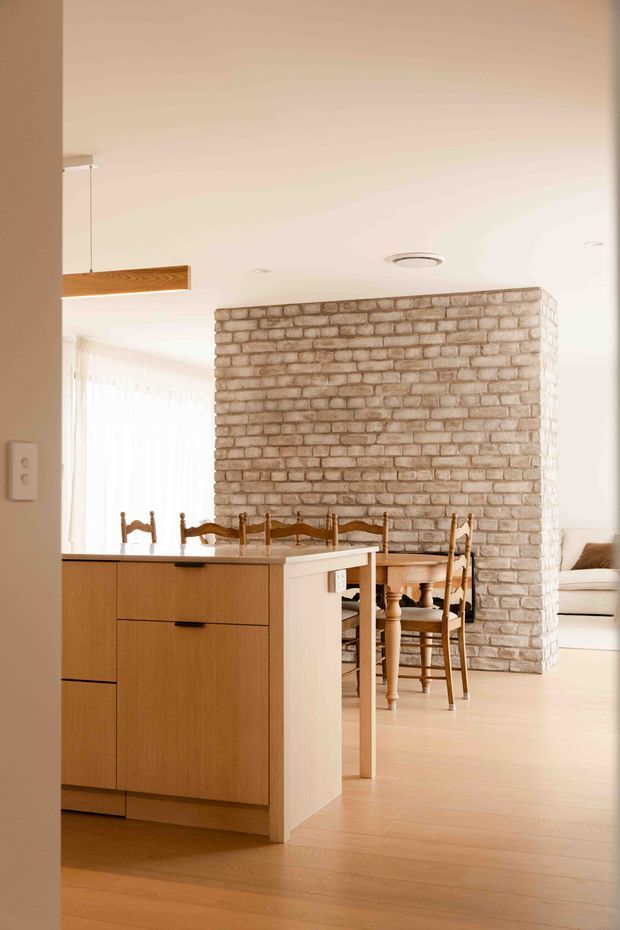 Bright open-plan residential interior viewed from an entry or hallway, featuring a compact timber kitchen island with dark benchtop, a dining area with traditional wooden chairs, pale timber flooring, and a prominent exposed light brick feature wall. A living area is partially visible beyond the dining space. The room has a warm, airy, contemporary-natural aesthetic with simple recessed lighting and a suspended timber light fixture.