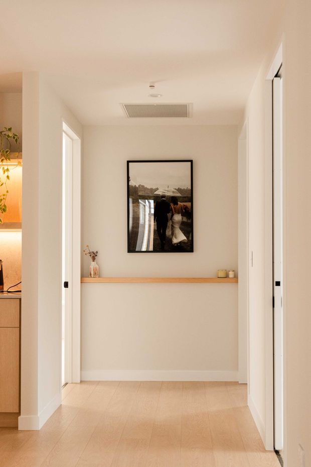 A bright, narrow residential hallway with warm pale timber flooring, off-white walls and trim, and a simple recessed timber display ledge. A framed photograph hangs centrally above the ledge, with small decorative objects placed along it. Open doorways reveal part of a warm-toned kitchen on the left and adjacent rooms on the right. The ceiling includes a rectangular air-conditioning vent and recessed fittings.