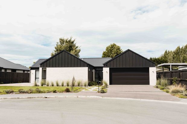 Street-facing view of a contemporary single-storey residence with two steeply gabled wings, dark vertical metal cladding and roofing, pale masonry walls, and a recessed double garage. The front garden uses low ornamental grasses and restrained planting, with a broad paved driveway and suburban street in the foreground.