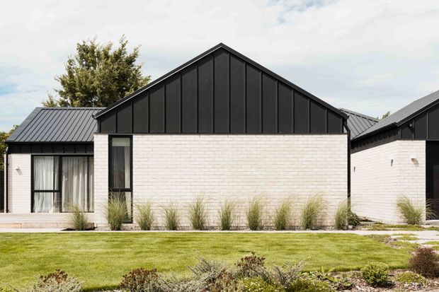 A contemporary single-storey residential exterior composed of connected gabled volumes. The main facade combines white brick masonry with a tall black standing-seam metal gable and dark metal roof, complemented by black-framed glazing and landscaped lawn planting.