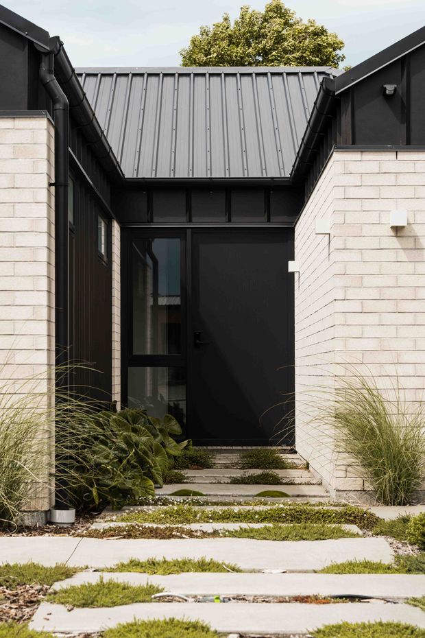 A contemporary residential entry viewed from the exterior, framed by pale brick walls and dark metal cladding. A black entry door with adjacent glazing sits beneath a standing-seam metal roof, with concrete stepping slabs, gravel, and layered ornamental planting leading toward the entrance.