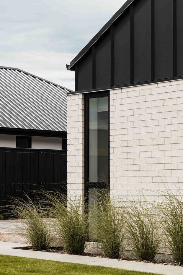 A close exterior architectural view of a contemporary building, showing pale white masonry walls, a dark upper facade with vertical paneling, a narrow black-framed window, and a corrugated metal roof on the adjoining structure. Ornamental grasses line a concrete pathway in the foreground.
