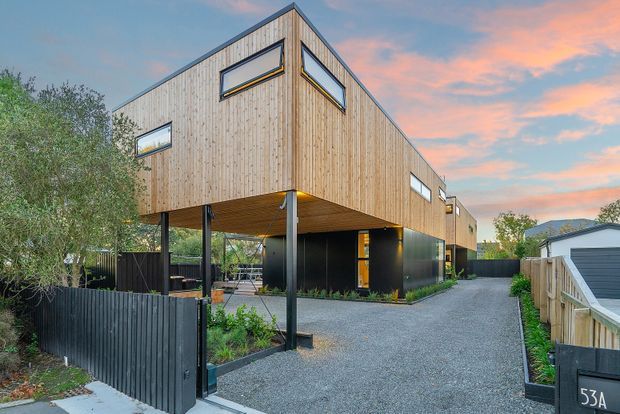 Exterior view of a contemporary elevated residence with a strong angular gable-like roof form, vertical timber cladding, dark lower-level walls, and an open undercroft/carport area supported by slender posts. Narrow horizontal windows punctuate the timber facade. A gravel driveway, dark fencing, neighbouring houses, and landscaping surround the building, shown in warm evening light.