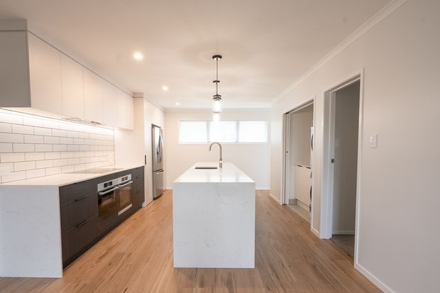 Bright contemporary kitchen with a long central island, white flat-panel cabinetry, dark integrated oven cabinetry, white subway-tile splashback, timber-look flooring, and pendant lighting. The room opens toward utility or adjoining spaces along the right wall and has a clean, neutral finish.