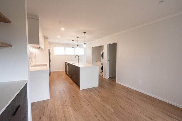 Bright open-plan kitchen and living area with warm timber flooring, white cabinetry, a large white island with an integrated sink, pendant lights, and minimal contemporary finishes. A laundry or utility area is visible through the doorway at the rear.