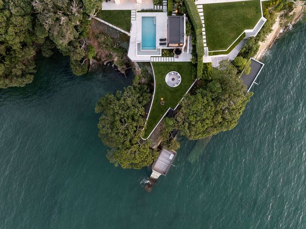 Aerial view of a landscaped waterfront property set on a wooded peninsula. The image shows a modern residence with a rectangular swimming pool, expansive lawn areas, curving paved paths, dense mature trees, and a small waterside jetty or deck surrounded by deep blue-green water.