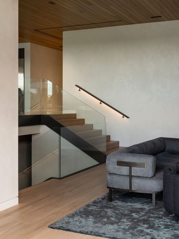Contemporary interior showing a timber-lined ceiling, pale plaster walls, warm timber flooring, and a sculptural stair with clear glass balustrades and a dark metal handrail. A dark grey sectional sofa and upholstered armchair occupy the foreground living area, with a textured rug beneath. The stair connects to a partially visible upper level and creates a strong architectural focal point.