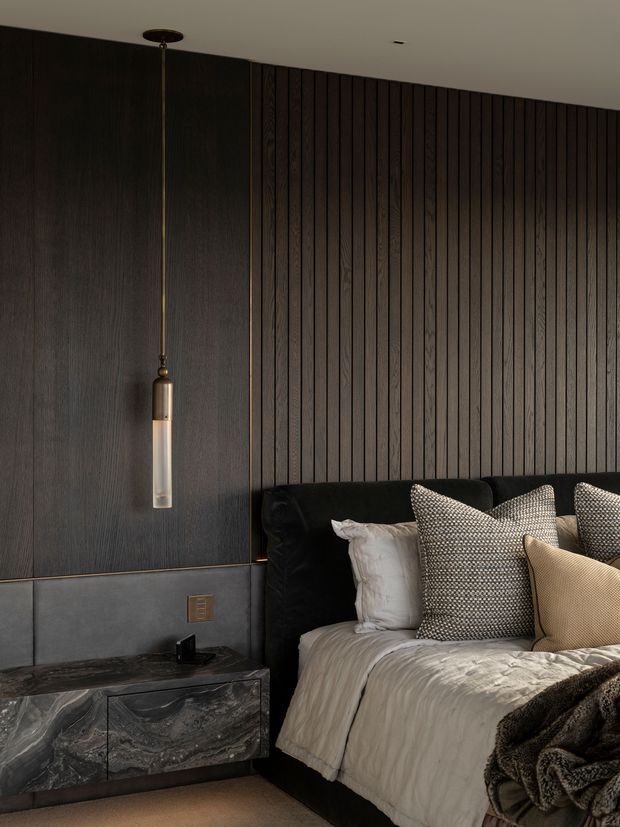 Moody contemporary bedroom with a dark upholstered bed, layered neutral bedding and textured cushions. The feature wall combines dark vertical timber battens with large dark timber panels, while a grey marble-look bedside cabinet and a slender pendant light add refined contrast. The room has a low, intimate feel and sophisticated dark neutral palette.
