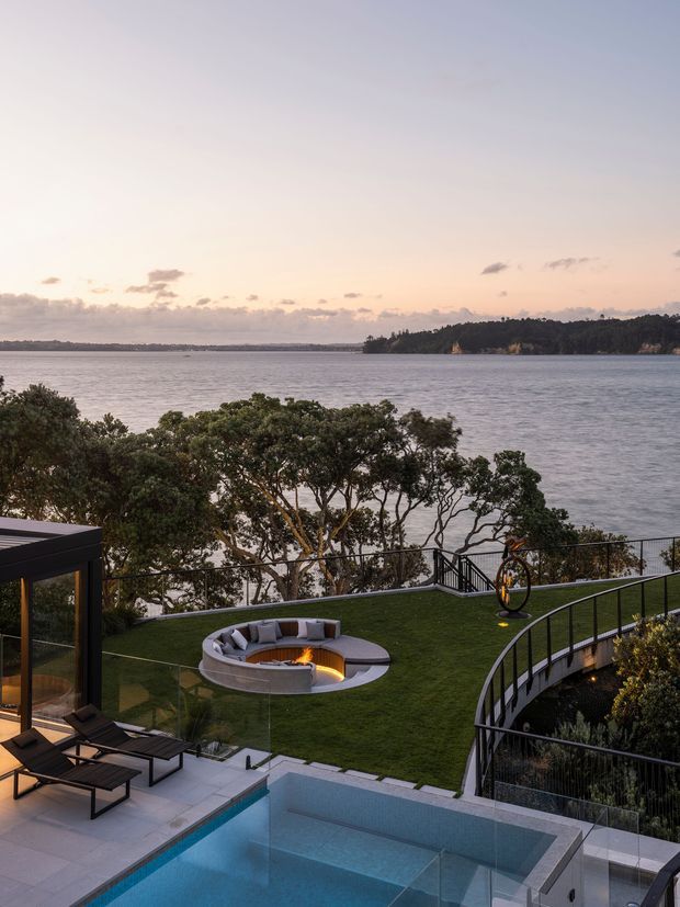 A landscaped waterfront residential outdoor area viewed from an elevated terrace, featuring a swimming pool, glass balustrades, a circular sunken fire pit with built-in seating, lounge chairs, manicured lawn, and curving stairways overlooking the water.