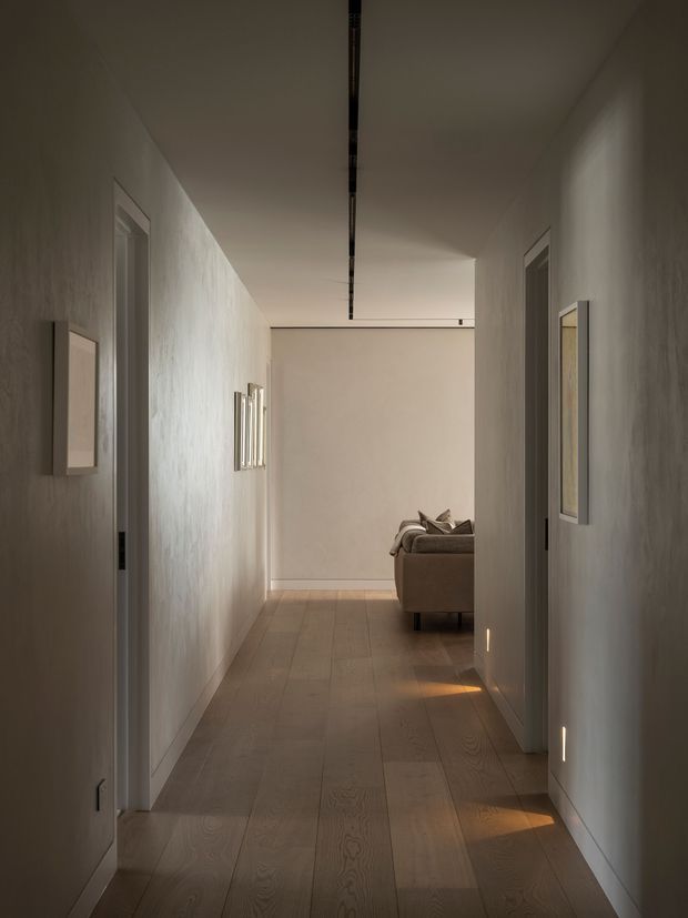 A long, narrow residential hallway with warm timber plank flooring, textured pale walls, flush doorways, framed wall artwork, and discreet linear ceiling and low-level wall lighting. A sofa is partially visible at the far end, suggesting an adjacent living room. The restrained palette and concealed lighting create a calm, refined contemporary interior.