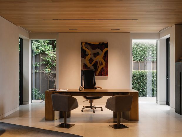 A refined home office with a large timber desk, two upholstered visitor chairs, and a central executive chair. The room features a warm timber slatted ceiling, pale neutral walls, light tiled flooring, full-height glazing and doors opening to landscaped greenery, with an abstract artwork above the desk and integrated linear ceiling lighting.