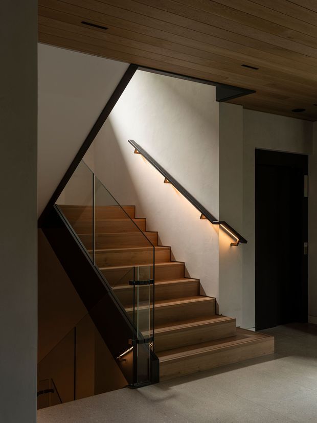 A refined contemporary interior stairway with warm timber treads and ceiling panelling, dark structural and handrail elements, clear glass balustrades, and integrated warm lighting along the wall-mounted handrail. The staircase rises beside a softly textured light wall, with a dark doorway visible at right and a polished neutral floor at the landing.