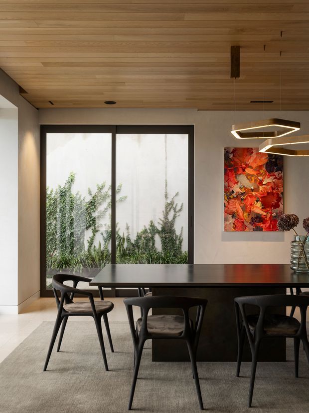 Contemporary dining area with a long dark dining table and sculptural black chairs, set beneath a warm timber-lined ceiling. Large black-framed glazed doors or windows open the room visually to a planted courtyard, while geometric pendant lights and a vivid abstract artwork add distinctive accents. Neutral plaster walls, pale flooring, and a large muted rug create a refined, warm interior.