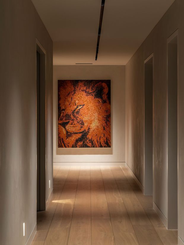 A narrow, refined residential hallway with warm beige plastered walls, light timber flooring, recessed doorways on both sides, and a linear recessed ceiling light. A large vivid orange and black artwork forms the focal point at the end of the corridor.