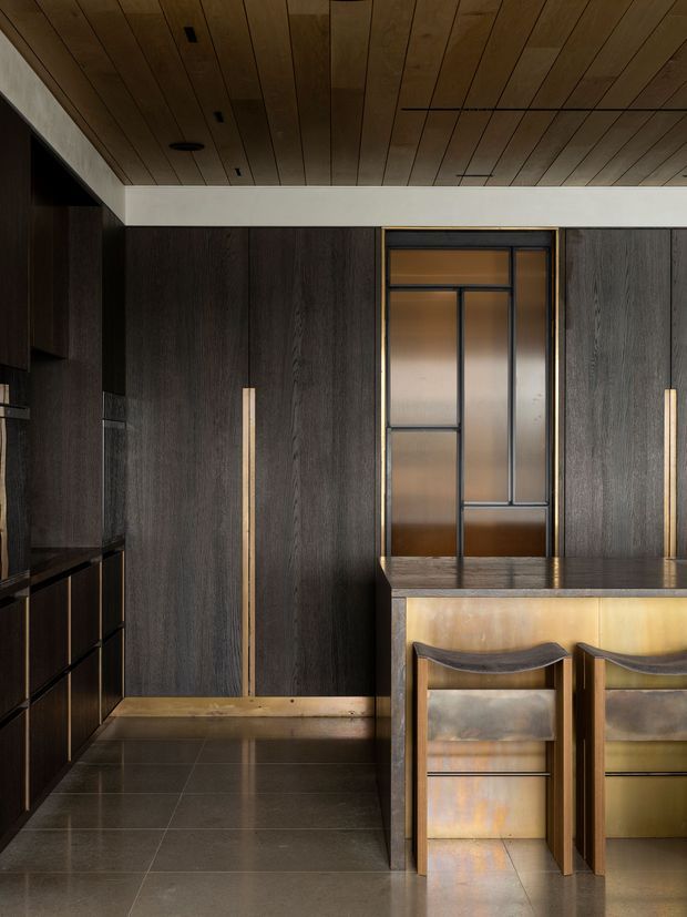 A refined, dark-toned kitchen with full-height charcoal wood cabinetry, integrated black appliances, a compact island with two stools, and a framed glazed door or partition at the rear. The ceiling is clad in warm timber boards, while the floor is finished in large dark tiles. Brass-toned trim and warm lighting add contrast to the moody, sophisticated interior.